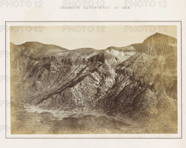 Tenerife Experiment of 1856, View of the crater of El Teide on Tenerife, photograph, page, number: 'Plate 3.', inscription: 'Portion of a Photograph (magnified 9.1 diameters), representing a mountain side (the interior S. Eastern wall of the Great Crater), four miles distant from the Camera, stationed at a height of 10, 700 feet above the level of the Sea. See pp 513, 514.', photographer: Charles Piazzi Smyth, Tenerife, printer (person): JA Spencer (possibly), 1856, printer (person), paper, albumen print, height 128 mm x width 197 mm, Photo in the section at the back., experiment, test, science and technology, volcano, A sepia-toned photograph of a rugged mountain landscape framed by a pale border, showing layered ridges and steep rocky slopes with scattered low vegetation, terraces and cliffs visible along the higher slopes, a broad valley floor with uneven ground and textured scrub, and distant rounded peaks beneath a pale sky.