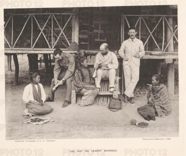 The way we learned Burmese, Three men with books and three women from Burma in front of a house, page, photomechanical print, photographer: AGE Newland, Burma, maker: Michel Berthaud, Paris, c. 1892, paper, collotype, photoprint, height 154 mm x width 202 mm, Prints opposite page 20., book, hut, cabin, lodge, A group of people sits and kneels in front of a raised wooden structure with a woven bamboo railing and vertical posts; two men in light-colored suits sit or stand near the railing, one seated reading from a small book and the other standing with a hand resting on the railing next to a hat, while another man in a darker uniform leans forward showing a piece of paper to a seated woman in patterned clothing who holds a small bundle in her lap; two other women kneel on the ground at either side, one with a scarf around her neck and a cloth of small round objects spread before her and the other wrapped in a patterned shawl holding a small item near her mouth with plates of small round...