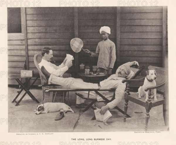 Two seated men and a boy with a fan behind them, The long, long Burmese day, page, photomechanical print, photographer: AGE Newland, Burma, maker: Michel Berthaud, Paris, c. 1892, paper, collotype, photoprint, height 157 mm x width 202 mm, dog, reading, fan, A monochrome scene shows a wooden paneled wall as a backdrop with three men and two dogs arranged on a porch. One man reclines in a wicker chair wearing light clothing and a turban, his legs stretched out and a pipe in his mouth while he reads a folded paper; another man reclines on a lounge with his head wrapped and eyes closed, one arm resting behind his head and the other hand holding a book; a standing man in a buttoned shirt and a turban holds a round woven fan on a handle over the seated men. A small round table between the chairs holds glassware and a covered jar. Two dogs are present, one lying on the floor near a shoe and the other sitting alert on a wooden chair beside the reclining man. The floor is covered with mats and...