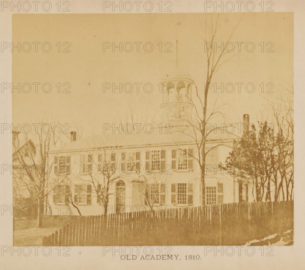 View of the first Friends Academy location in New Bedford, Massachusetts, Old Academy. 1810, photograph, page, c. 1871 - in or before 1876, photographer, paper, albumen print, height 109 mm x width 132 mm, scholastic education, tuition, civic architecture, edifices, dwellings, North America, New Bedford, A faded sepia photograph of a large rectangular building with multiple shuttered windows across two stories, a pitched roof, and a small central doorway, partially obscured by leafless trees in front and a wooden picket fence along the foreground; a cupola with arched openings rises above the roofline and a tall thin pole extends upward nearby, while more trees and a chimney appear on either side, all shown against a pale sky.