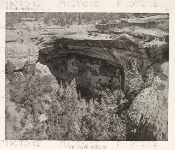 The Cliff Palace, View of the Cliff Palace in Mesa Verde National Park, page, photomechanical print, number: 'XII', inscription: 'G. Nordenskiöld: Klippboningar.', photographer: Gustaf Nordenskiöld, Mesa Verde National Park, cliché maker: Librairies-Imprimeries Réunies (Ancienne Maison Quantin), Paris, c. 1891 - in or before 1893, North America, Colorado, paper, photoaquatint, height 190 mm x width 242 mm, ruin of a dwelling, house, castle, cave-dwelling, Cliff Palace, A wide rock overhang forms a recessed alcove in a cliff face, with layered rock strata and textured surfaces visible on the ceiling and walls of the overhang; beneath the shelter, a cluster of multi-level stone structures with rectangular openings and angular walls sits among scattered boulders and rubble; the foreground shows a slope of rocky terrain and dense vegetation with leafy and twiggy shrubs and small trees, some with pale, bare branches; the upper area of the scene displays a rough plateau of rock with sparse l...