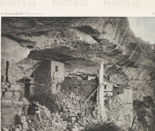 The Balcony House, Balcony House in Mesa Verde National Park, page, photomechanical print, number: 'XIV', inscription: 'G. Nordenskiöld: Klippboningar.', photographer: Gustaf Nordenskiöld, Mesa Verde National Park, cliché maker: Librairies-Imprimeries Réunies (Ancienne Maison Quantin), Paris, c. 1891 - in or before 1893, North America, Colorado, paper, photoaquatint, height 189 mm x width 240 mm, ruin of a dwelling, house, castle, cave-dwelling, A black and white photograph showing a rocky cliff overhang with rough textured rock surfaces and striations, beneath which are multiple stone structures in varying states of ruin with partial walls, narrow rectangular openings, and a freestanding chimney built of stacked stone, piles of loose stones and rubble scattered around, wooden beams and poles leaning against walls and among the debris, sparse vegetation and small trees visible near the cliff edge, and a seated person wearing a hat and darker clothing holding an object while sitting on ...