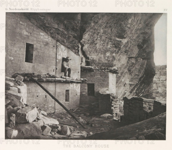 The Balcony House, Balcony House in Mesa Verde National Park with a man in a frame, page, photomechanical print, inscription: 'G. Nordenskiöld: Klippboningar.', number: 'XV', photographer: Gustaf Nordenskiöld, Mesa Verde National Park, cliché maker: Librairies-Imprimeries Réunies (Ancienne Maison Quantin), Paris, c. 1891 - in or before 1893, North America, Colorado, paper, photoaquatint, height 190 mm x width 240 mm, ruin of a dwelling, house, castle, cave-dwelling, A weathered stone and plaster structure built against a large rock overhang with uneven rocky surfaces; a person wearing a wide-brimmed hat and light-colored clothing sits in a doorway on an upper ledge above a rough wooden beam and layered roof materials; rectangular openings and small windows punctuate the building walls, and stacked stones and rubble form low walls and piles on the ground in front; vertical and diagonal wooden supports lean against the structure, and the rocky cliff rises steeply behind and above the sce...