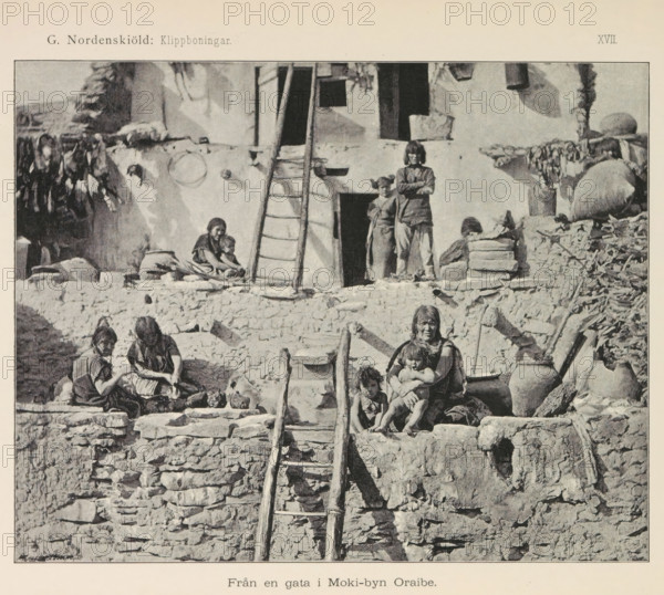 View of a village of the Hopi people, Moki-byn Wol'-pi, page, photomechanical print, name, number: 'Fig. 153.', photographer: Gustaf Nordenskiöld (possibly), North and Central America, cliché maker: Angerer & Göschl, C., Vienna, c. 1891 - in or before 1893, Gustaf Nordenskiöld, Angerer & Göschl, photographer, cliché maker, Lower Austria, paper, autotypie, height 90 mm x width 148 mm, prospect of village, silhouette of village, A grayscale photograph of a stone and plaster building with rough stacked stone walls and a wooden ladder leading up to a doorway, several people sitting and standing on the terraces and steps, a woman holding a child on a ledge with another child nearby, two women seated and working with items in a basin, two figures standing in a doorway on the upper level, pottery jars and bundles placed on the ledges, hanging items draped over the walls, and irregular stone steps and ledges forming the exterior façade.
