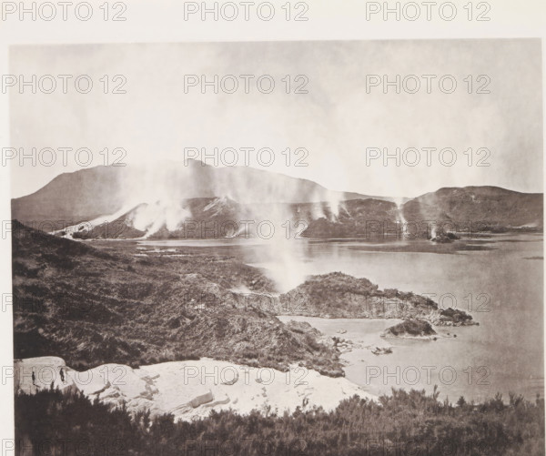 View of Lake Rotomahana and the Pink Terraces, page, photomechanical print, photographer: Daniel Louis Mundy, New Zealand, cliché maker: anonymous, United Kingdom, c. 1865 - c. 1875, paper, collotype, autotype, height 197 mm x width 246 mm, lake, hot spring, geyser, Lake Rotomahana, A wide landscape photograph showing a lake bordered by rocky shorelines and low hills, with steam or smoke rising in several narrow plumes from the ground along the distant shoreline and hills; the foreground includes light-colored rocky outcrops and darker vegetation, and two small human figures standing on a pale rock ledge near the lower left, with distant mountains forming a hazy horizon under a pale sky.