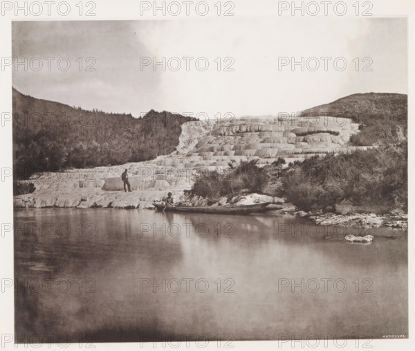View of the Pink Terraces along Lake Rotomahana, page, photomechanical print, photographer: Daniel Louis Mundy, New Zealand, cliché maker: anonymous, United Kingdom, c. 1865 - c. 1875, paper, collotype, autotype, height 202 mm x width 249 mm, lake, hot spring, geyser, steam, Lake Rotomahana, Pink Terraces, A wide monochrome landscape shows a calm reflective body of water in the foreground with a stepped white terraced formation rising behind it and continuing across the middle of the image, textured with horizontal ledges and small ridges; a wooded hillside frames the terraces on the left and right with darker trees and scrub, and a person stands on one of the lower terraces near the water while another person sits in a small boat at the water’s edge; the sky above is pale with soft cloud patterns and the overall scene has a vintage photographic tone.