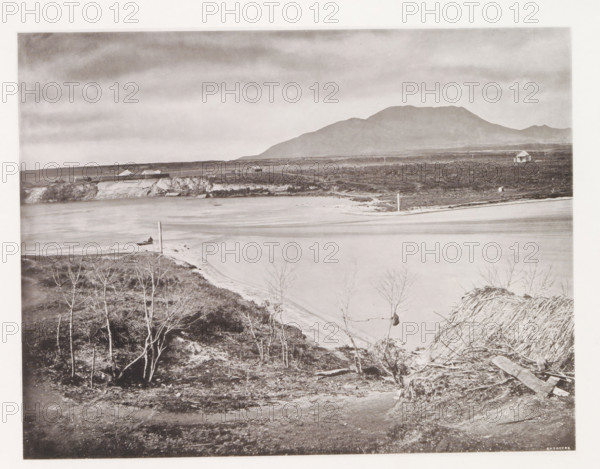 View of Lake Taupo, page, photomechanical print, photographer: Daniel Louis Mundy, New Zealand, cliché maker: anonymous, United Kingdom, c. 1865 - c. 1875, paper, collotype, autotype, height 192 mm x width 250 mm, lake, volcano, hot spring, geyser, Lake Taupo, A wide grayscale landscape showing a calm body of water curving along a sandy shore with a small boat near the waterline, low rocky cliffs and flat terrain behind the shore, scattered leafless trees and rough vegetation in the foreground, a few simple buildings and a solitary small house on the distant plain, telephone or utility poles dotting the scene, and a large rounded mountain range under a cloudy sky.