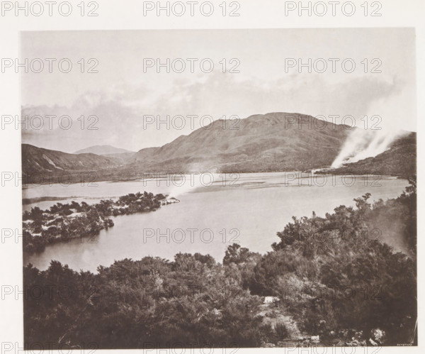 View of Lake Rotomahana, page, photomechanical print, photographer: Daniel Louis Mundy, New Zealand, cliché maker: anonymous, United Kingdom, c. 1865 - c. 1875, paper, collotype, autotype, height 201 mm x width 248 mm, lake, hot spring, geyser, Lake Rotomahana, A wide view of a lake bordered by shoreline vegetation in the foreground and an island with low trees extending into the water, with a range of rounded hills rising behind the lake and a plume of steam or smoke emerging from a rocky area on the right side, under a sky with light cloud coverage.