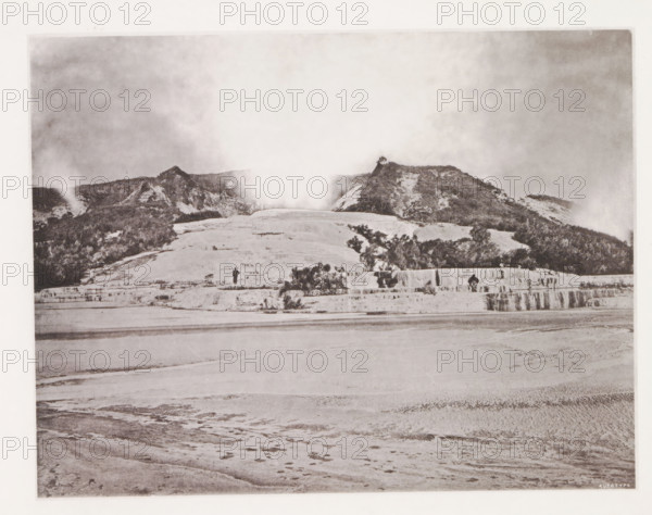 View of the White Terraces at Lake Rotomahana, page, photomechanical print, photographer: Daniel Louis Mundy, New Zealand, cliché maker: anonymous, United Kingdom, c. 1865 - c. 1875, paper, collotype, autotype, height 198 mm x width 245 mm, lake, hot spring, geyser, Lake Rotomahana, White Terraces, A wide sepia-toned landscape showing a layered mineral terrace in the foreground with smooth, rippled surfaces and shallow pools, rising to a stepped shelf where shrubs and small bushes grow, beyond which a rounded slope leads up to a ridge of rocky hills covered with sparse vegetation and patches of lighter exposed rock, a plume of steam or mist rising from the center of the ridge and drifting into a cloudy sky above, and two small human figures standing on the middle terrace near the vegetation.