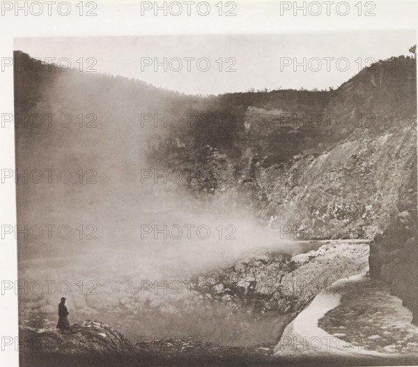 View of a geyser in Lake Rotomahana, page, photomechanical print, photographer: Daniel Louis Mundy, New Zealand, cliché maker: anonymous, United Kingdom, c. 1865 - c. 1875, paper, collotype, autotype, height 208 mm x width 250 mm, lake, hot spring, geyser, Lake Rotomahana, A sepia-toned scene showing a rocky cliffside and a steep ravine with mist or fog drifting across the middle of the image, a lone silhouetted figure standing on a rocky outcrop at the lower left edge, rough textured rock surfaces and a narrow winding path or ledge along the right edge, distant tree-lined ridgelines visible along the top, and variations in light and shadow creating a hazy atmosphere.
