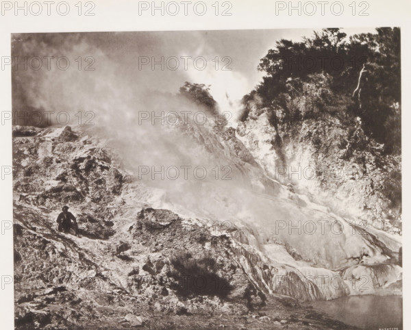 Geyser near Lake Rotomahana, page, photomechanical print, photographer: Daniel Louis Mundy, New Zealand, cliché maker: anonymous, United Kingdom, c. 1865 - c. 1875, paper, collotype, autotype, height 198 mm x width 253 mm, lake, hot spring, geyser, steam, Lake Rotomahana, A black-and-white photograph showing a rocky landscape with steam or mist rising from the ground and flowing across the scene, rough textured terraces and mineral deposits forming layered slopes and ledges, a darker forested area with dense trees and vegetation on the upper right, a solitary figure wearing a hat and dark clothing seated on a rocky outcrop near the lower left, and a pool of water partially visible at the lower right edge.