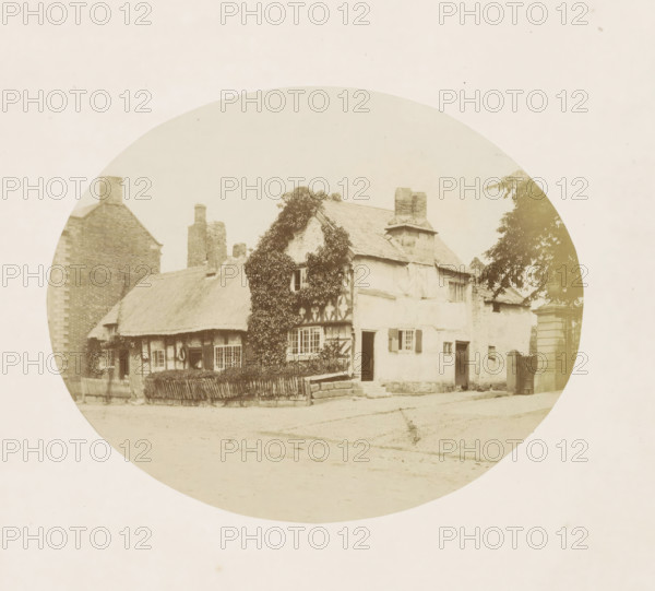 View of a house in Tarvin, Cottage in Tarvin, photograph, page, photographer: William Bryans, Tarvin, c. 1850 - in or before 1858, Cheshire (county), paper, albumen print, height 141 mm x width 175 mm, cottage, A circular sepia photograph shows an old cottage with a thatched roof and timber-framed walls, part of the facade covered in climbing ivy, multiple windows with small panes and shutters, an open doorway, a low wooden fence in front, a stone wall and steps leading up to the entrance, a tall brick chimney and additional brick buildings adjacent, a tree partially visible on one side, and an unpaved road or courtyard in the foreground.