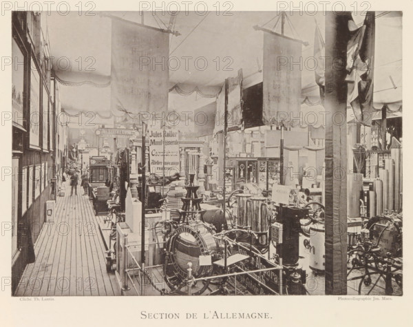 Section de l'Allemagne, Exhibition of German objects during the world exhibition in Antwerp, page, photomechanical print, photographer: Th. Lantin, Antwerp, cliché maker: Joseph Maes, 1894, Th. Lantin, Joseph Maes, photographer, cliché maker, paper, collotype, light printing, height 160 mm x width 220 mm, world fair, world exhibition, exhibition, show, A long indoor exhibition aisle with wooden plank flooring bordered by booths and displays; hanging fabric banners drape from the ceiling and decorative scalloped valances run along the upper walls; rows of framed pictures line the left wall while a variety of machinery, metal equipment, stacked parts, and display cabinets fill the central and right floor space; tall vertical posts and partitions separate sections and a few indistinct human figures are visible near the far end of the aisle; the overall image has a sepia tone and a high vantage capturing depth down the corridor.