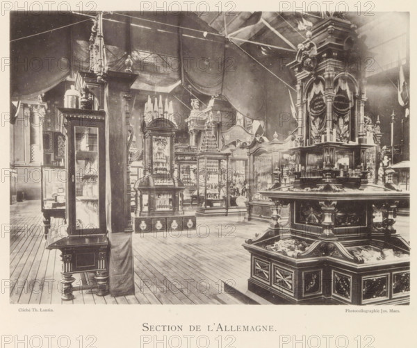 Exhibition of German objects during the world exhibition in Antwerp, Section de l'Allemagne, page, photomechanical print, photographer: Th. Lantin, Antwerp, cliché maker: Joseph Maes, 1894, Th. Lantin, Joseph Maes, photographer, cliché maker, paper, collotype, light printing, height 161 mm x width 218 mm, world fair, world exhibition, exhibition, show, A sepia-toned interior scene showing a large exhibition room with wooden plank flooring and high draped fabric hanging from the ceiling, filled with ornate display cabinets and sculptural wooden fixtures. Prominent carved wooden pedestals and glass-fronted cases stand throughout the space, many with decorative moldings, columns, and finials. A large multi-tiered central display with turned legs and carved panels occupies the foreground, its upper surface holding small objects and decorative elements. In the midground several tall glass vitrines showcase grouped items and have elaborate crowned tops, while a rounded cabinet with a domed t...