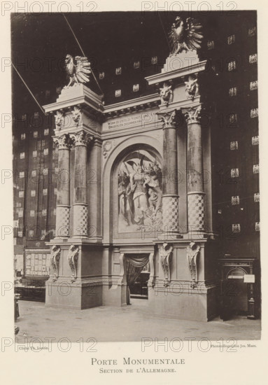 Porte Monumentale Section de l'Allemagne, Gate in an exhibition of German objects during the world exhibition in Antwerp, page, photomechanical print, photographer: Th. Lantin, Antwerp, cliché maker: Joseph Maes, 1894, Th. Lantin, Joseph Maes, photographer, cliché maker, paper, collotype, light printing, height 215 mm x width 160 mm, gate, entrance, world fair, world exhibition, exhibition, show, A large ornate stone gateway occupies the image, featuring tall fluted columns with decorated capitals and patterned lower shafts, sculpted lion heads above the entablature, and perched eagle sculptures on the upper corners; a large arched recess in the central section contains a high-relief sculptural group of robed figures and foliage, and a draped fabric hangs in the opening beneath the arch; the surrounding wall shows a repeated decorative motif, and furniture and architectural details are visible at floor level beside the gateway.