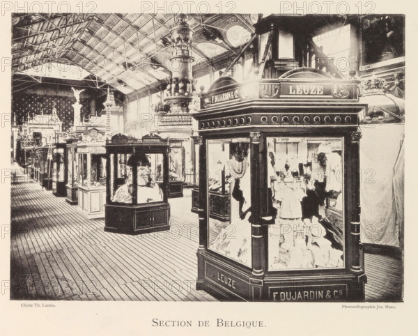 Section de Belgique, Expositie van Belgische beelden tijdens de wereldtentoonstelling te Antwerpen, page, photomechanical print, photographer: Th. Lantin, Antwerp, clichémaker: Joseph Maes, 1894, paper, collotype, light print, height 160 mm x width 220 mm, world fair, world exhibition, exhibition, show, A large indoor exhibition space with a high vaulted ceiling supported by visible metal trusses and skylights, filled with ornate display kiosks and glass cases showcasing garments and decorative objects, a wooden plank floor running parallel to the display rows, an elaborately carved central column with layered decorative elements rising toward the ceiling, arched windows along the far wall letting in light, sculptural figures and ornamental architectural details visible behind and between the kiosks, and various draped fabrics and mannequins positioned within the front glass case.