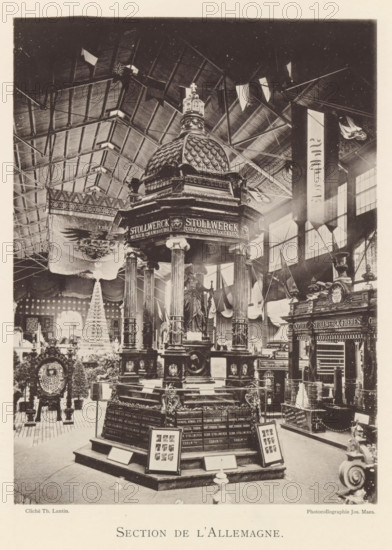 Section de l'Allemagne, Exhibition of the Stollwerck company at the World Exhibition in Antwerp, page, photomechanical print, photographer: Th. Lantin, Antwerp, cliché maker: Joseph Maes, 1894, Th. Lantin, Joseph Maes, photographer, cliché maker, paper, collotype, light printing, height 215 mm x width 160 mm, world fair, world exhibition, exhibition, show, A large indoor exhibition scene with a tall ornate pavilion topped by a domed roof and crowned finial, supported by fluted columns and decorative pedestals, surrounded by display cases and ornamental objects. The interior has a high vaulted ceiling with visible trusses and hanging pennants, and tall windows along one wall letting in light. Flags and banners hang from the ceiling and walls. Decorative sculptures and relief elements adorn the pavilion and nearby structures, and framed panels and small display stands are arranged around the base. In the background a tiered, cone-shaped decorative installation and patterned draperies are...