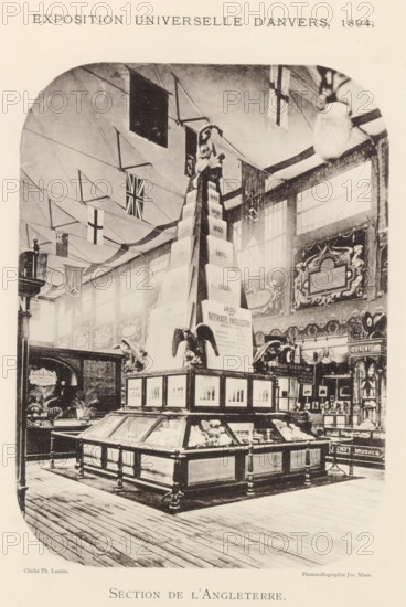 Exhibition of British objects during the World Exhibition in Antwerp, Section de l'Angleterre, page, photomechanical print, photographer: Th. Lantin, Antwerp, clichémaker: Joseph Maes, 1894, Th. Lantin, Joseph Maes, photographer, clichémaker, paper, collotype, photoprint, height 220 mm x width 160 mm, world fair, world exhibition, exhibition, show, A large indoor exhibition hall with high windows and decorative wall panels, featuring a central tall pyramid-like display topped by a seated female figure and flanked by sculpted eagle figures; glass-topped wooden display cases surround the base of the central structure, containing objects and framed panels on their sides; flags and bunting hang from the ceiling across the space; ornate signage and decorative motifs adorn the walls behind the displays; wooden floorboards run across the foreground.