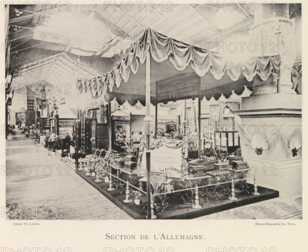 Exhibition of German objects during the world exhibition in Antwerp, Section de l'Allemagne, page, photomechanical print, photographer: Th. Lantin, Antwerp, cliché maker: Joseph Maes, 1894, Th. Lantin, Joseph Maes, photographer, cliché maker, paper, collotype, photoprint, height 158 mm x width 220 mm, world fair, world exhibition, exhibition, show, A large indoor exhibition space with a high vaulted roof and visible metal trusses, rows of display booths and ornate decorative columns, a central raised platform area enclosed by rope and stanchions holding various ornate machinery and wooden cabinets, a fabric canopy with scalloped and fringed drapery suspended over the central area supported by slender poles, decorative plants placed around the displays, a person seated near the front of the exhibit partially visible behind equipment, additional booths and objects extending into the distance along a wooden plank floor, and decorative architectural elements including carved column bases a...