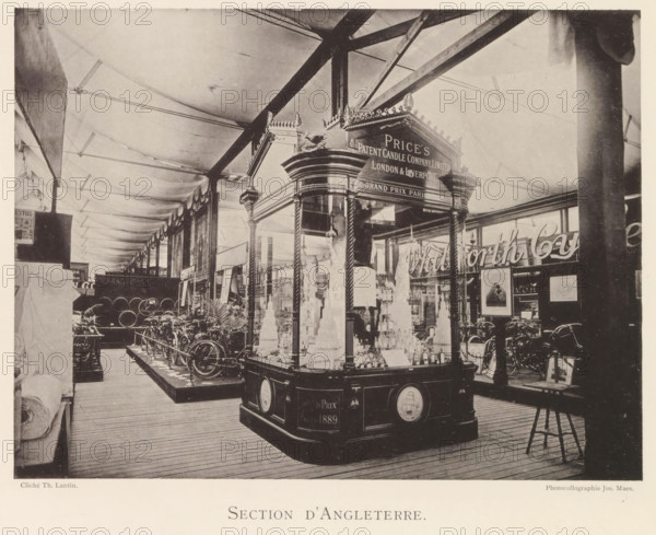 Section d'Angleterre, Exhibition of British objects during the World Exhibition in Antwerp, page, photomechanical print, photographer: Th. Lantin, Antwerp, clichémaker: Joseph Maes, 1894, Th. Lantin, Joseph Maes, photographer, clichémaker, paper, collotype, photoprint, height 160 mm x width 214 mm, world fair, world exhibition, exhibition, show, A large indoor exhibition space with a high fabric ceiling supported by visible beams and columns, showing a decorative wooden display pavilion with glass panels and ornate trim in the foreground filled with tiered objects and bottles, flanked by rope-twisted columns and a carved cornice topped by ornamental finials. Around the pavilion are rows of bicycles and metal wheels arranged on raised platforms, various framed pictures and signs mounted on walls and stands, potted plants near the floor, wooden plank flooring underfoot, and a tall dark column partially obscuring the right side of the scene. The image has a sepia tone and shows detailed s...