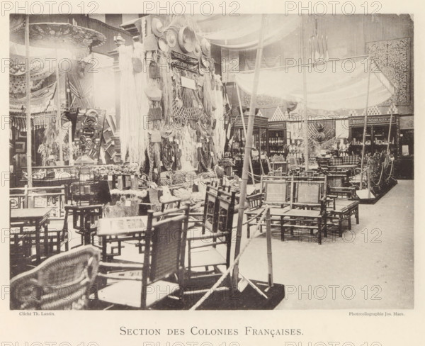 Section des Colonies Françaises, Exhibition of the French colonies during the World Exhibition in Antwerp, page, photomechanical print, photographer: Th. Lantin, Antwerp, cliché maker: Joseph Maes, 1894, paper, collotype, photoprint, height 160 mm x width 219 mm, colonial system, world fair, world exhibition, exhibition, show, A large indoor scene showing many wooden chairs arranged in rows and stacks across the foreground and midground, woven seats and backs visible on several pieces, with a variety of baskets, ropes, and hanging textile fringes displayed on a central vertical structure. Canopy fabrics and draped cloth hang from poles overhead, and decorative plates and woven mats are mounted on the display. Shelving units and display cabinets filled with bottles and containers sit toward the rear, flanked by patterned wall hangings and carved panels. Several poles and ropes run through the space, and small tables, stools, and ornamental objects are scattered among the furniture and d...