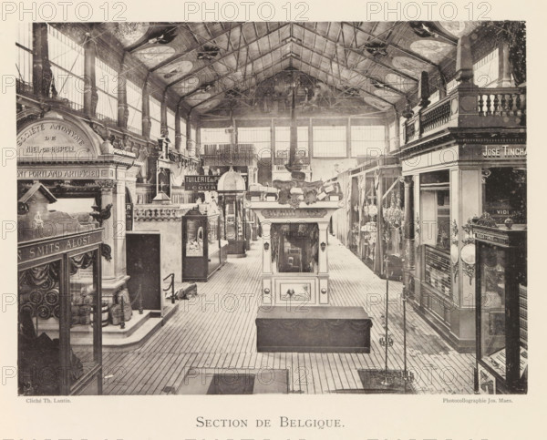 Section de Belgique, Expositie van Belgische beelden tijdens de wereldtentoonstelling te Antwerpen, page, photomechanical print, photographer: Th. Lantin, Antwerp, clichémaker: Joseph Maes, 1894, paper, collotype, light print, height 160 mm x width 219 mm, world fair, world exhibition, exhibition, show, A wide indoor hall with a high vaulted ceiling supported by visible metal trusses and decorated panels, tall windows along both side walls letting in light, a central aisle with wooden plank flooring flanked by display booths and ornate architectural facades, a raised decorative kiosk in the middle foreground with sculptural elements and framed panels, various display cases and shelving units containing objects, carved columns and balustrades atop some booths, hanging chandeliers and lamps along the right side, ornamental moldings and statues integrated into the booths, and smaller decorative items and furnishings arranged throughout the space.