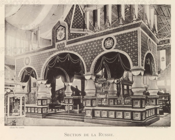 Section de la Russie, Exhibition of Russian objects during the World Exhibition in Antwerp, page, photomechanical print, photographer: Th. Lantin, Antwerp, clichémaker: Joseph Maes, 1894, paper, collotype, photoprint, height 160 mm x width 219 mm, world fair, world exhibition, exhibition, show, A large ornate pavilion with patterned walls and decorative arches stands inside a spacious hall; the structure features rounded archways draped with fabric swags, heavy column-like supports with layered bases and capitals, and panels with repeating motifs covering the upper surfaces. Above the arches a peaked roof section with patterned surfaces and circular medallions bearing emblem-like designs rises toward the ceiling, where hanging fabric banners descend from an exposed truss roof. In front of and beneath the arches are display areas and small stands with varied objects, and a framed glass cabinet sits at one side showing items inside. The scene is monochrome and shows varied textures of ca...