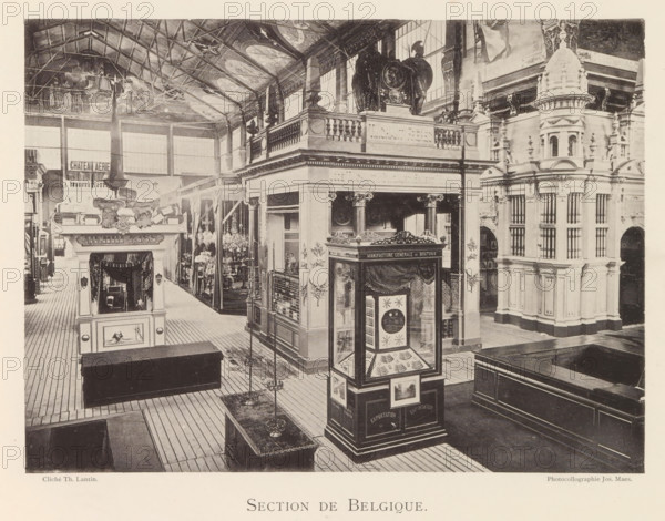 Section de Belgique, Expositie van Belgische beelden tijdens de wereldtentoonstelling te Antwerpen, page, photomechanical print, photographer: Th. Lantin, Antwerp, clichémaker: Joseph Maes, 1894, paper, collotype, light print, height 160 mm x width 220 mm, world fair, world exhibition, exhibition, show, A sepia-toned photograph showing an indoor exhibition hall with high vaulted ceiling and visible rafters, a wide wooden plank floor, and tall windows along the back wall; ornate multi-level architectural displays and pavilions fill the space, featuring classical columns, balustrades, carved decorative elements, and a domed turret-like structure on the right; a freestanding glass display case with decorative panels and framed images stands near the foreground, and a smaller kiosk with sculpted ornamentation and draped fabric is visible toward the left; hanging chandeliers and tall lampstands are arranged along the center aisle, and carved statues and reliefs decorate the upper terraces a...