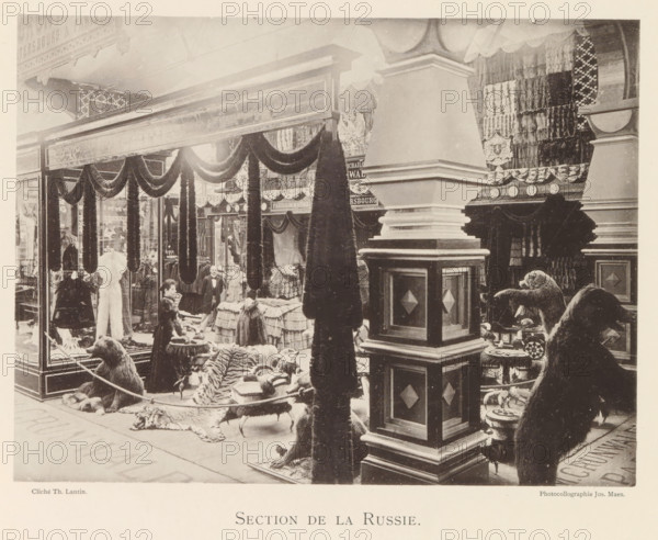 Section de la Russie, Exhibition of Russian fur products during the World Exhibition in Antwerp, page, photomechanical print, photographer: Th. Lantin, Antwerp, clichémaker: Joseph Maes, 1894, paper, collotype, photogravure, height 163 mm x width 219 mm, world fair, world exhibition, exhibition, show, furrier, A large indoor display area with tall decorative columns and draped fabric swags framing a storefront scene, shelves and racks filled with folded textiles and fur garments, several standing fur coats and hanging pelts, a woman near a counter arranging items, other figures standing behind tables covered in stacked goods, rugs and animal hides spread on the floor including a striped hide, mounted taxidermy bears positioned near stools and low tables, ornate carved wood and paneling on the pillars, patterned wall hangings and shields on the back walls, and a glass display case at the left showing garments inside.