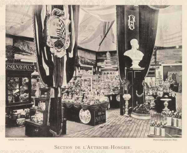 Exhibition of Austro-Hungarian ceramics during the world exhibition in Antwerp, Section de l'Autriche-Hongrie, page, photomechanical print, photographer: Th. Lantin, Antwerp, cliché maker: Joseph Maes, 1894, Th. Lantin, Joseph Maes, photographer, cliché maker, paper, collotype, photoprint, height 165 mm x width 218 mm, container of ceramics: jar, jug, pot, vase, world fair, world exhibition, exhibition, show, A large indoor display area filled with ceramics, vases, plates, figurines, and decorative objects arranged on tables and shelves beneath draped curtains and banners; a sculpted bust sits on a tall pedestal near the center-right, flanked by tall ornamental columns and urns, while ornate framed shields and patterned fabrics hang from the curtains; wooden floorboards run across the foreground and bottles and stacked containers occupy a small platform on the right; in the background are painted scene backdrops including a statue on a pedestal and architectural elements, with many sma...