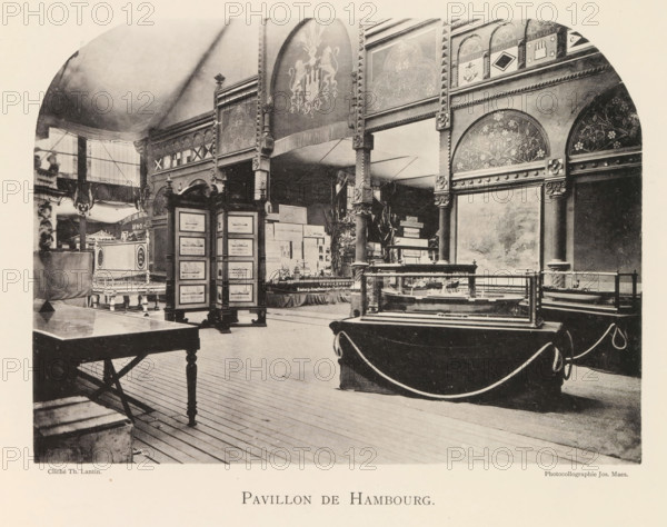 Interior of the pavilion from Hamburg during the World Exhibition in Antwerp, Pavillon de Hambourg, page, photomechanical print, photographer: Th. Lantin, Antwerp, clichémaker: Joseph Maes, 1894, paper, collotype, photogravure, height 160 mm x width 219 mm, interior, representation of a building, world fair, world exhibition, exhibition, show, A large interior space with a high curved ceiling and patterned decorative panels along the upper walls, featuring ornate arches and floral motifs. Wooden columns and carved supports rise from the floor to the decorated frieze, with a hanging fringed banner bearing a heraldic emblem. Display cases with glass tops sit on draped tables and pedestals, and a freestanding multi-paneled screen shows framed images. A wooden table with turned legs occupies the foreground, and ropes form a barrier around some exhibits. In the left background there is a raised platform with additional decorative furnishings and a tall window letting in light.