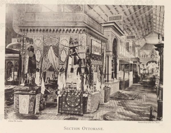 Exhibition of Ottoman objects during the World Exhibition in Antwerp, Section Ottomane, page, photomechanical print, photographer: Th. Lantin, Antwerp, clichémaker: Joseph Maes, 1894, paper, collotype, photoprint, height 160 mm x width 218 mm, interior, representation of a building, world fair, world exhibition, exhibition, show, A crowded indoor market aisle filled with decorative textiles and hanging fabrics displayed on stalls and mannequins, ornate carved and tiled architectural features including columns and an arched doorway, patterned rugs and embroidered cloths draped over tables and boxes, shelves and cases holding small objects, a wooden plank floor with rugs placed on it, a high glass and metal roof structure with hanging signs, and various decorative elements and merchandise arranged along the passage.