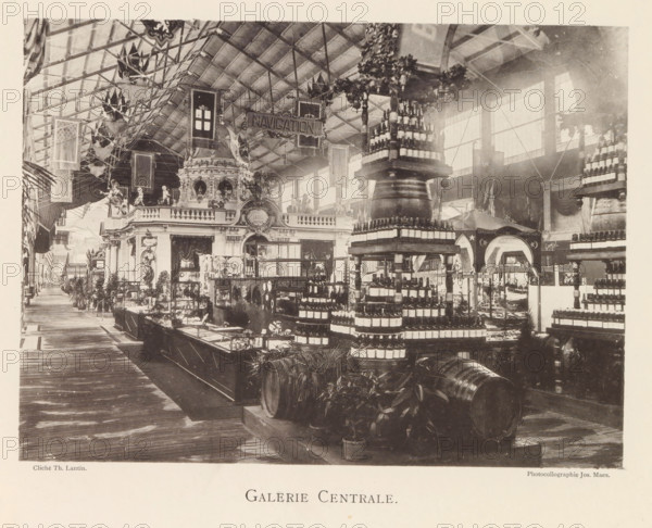 Interior of the central hall of the world exhibition in Antwerp, Galerie Centrale, page, photomechanical print, photographer: Th. Lantin, Antwerp, cliché maker: Joseph Maes, 1894, paper, collotype, light printing, height 160 mm x width 218 mm, interior, representation of a building, world fair, world exhibition, exhibition, show, A large indoor exhibition hall with a high vaulted ceiling supported by a visible metal framework and large windows along one side, filled with ornate displays and decorative architectural elements including a multi-level pavilion with sculpted figures and balustrades, rows of stacked bottles arranged on circular tiered stands and shelving, wooden barrels and potted plants positioned on the floor, a wide wooden walkway running alongside the exhibits, hanging banners and flags suspended from the ceiling, and various smaller display cases and counters spread throughout the space.