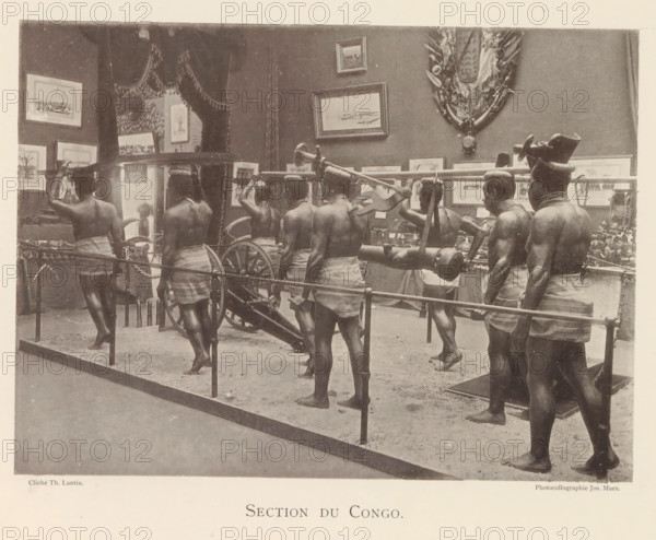 Sculptures in the Congolese part of the world exhibition in Antwerp, Section du Congo, page, photomechanical print, photographer: Th. Lantin, Antwerp, cliché maker: Joseph Maes, 1894, paper, collotype, photoprint, height 160 mm x width 219 mm, sculpture, world fair, world exhibition, exhibition, show, A sepia-toned scene showing a museum-like display of several bare-chested men wearing wrapped cloths around their waists and standing barefoot on a sandy floor, carrying a long pole with a mounted cylindrical object and wagon wheel parts, with framed paintings and decorative shields hung on the wall behind them, a roped barrier in front of the display, and a few additional artifacts and small objects arranged on tables along the back wall.