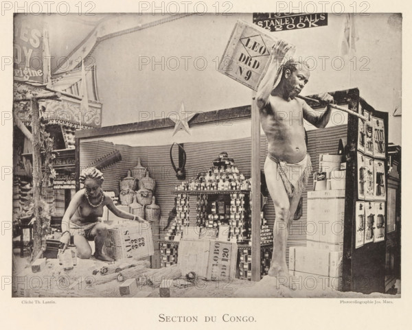 Sculptures in the Congolese part of the world exhibition in Antwerp, Section du Congo, page, photomechanical print, photographer: Th. Lantin, Antwerp, cliché maker: Joseph Maes, 1894, paper, collotype, photoprint, height 160 mm x width 218 mm, sculpture, world fair, world exhibition, exhibition, show, A sepia-toned photograph showing two partially clothed individuals posed among stacked boxes, jars, woven baskets, and various packaged goods on a display area framed by decorative panels and a star ornament; a seated figure wearing a beaded necklace and a cloth around the waist is reaching toward containers on the ground while a standing figure balanced on one leg holds a wooden pole across the shoulders and a crate over one shoulder; behind them are shelves filled with stacked jars and cylindrical containers, bundled brooms or reeds on the floor, and a tall display cabinet with panels showing relief images.