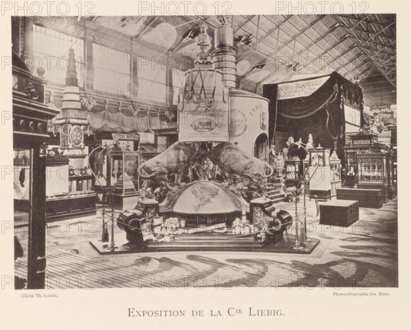 Exhibition of Liebig Meat Extracts during the World Exhibition in Antwerp, Exposition de la Cie Liebig, page, photomechanical print, photographer: Th. Lantin, Antwerp, clichémaker: Joseph Maes, 1894, paper, collotype, photogravure, height 144 mm x width 204 mm, foodstuffs, still life of foodstuffs, world fair, world exhibition, exhibition, show, A large indoor exhibition space with a high vaulted ceiling and tall windows shows an ornate central display featuring two large sculpted bulls positioned on a rounded base in front of a tall cylindrical column with decorative bands and medallions, topped by a bust. Surrounding the central piece are display cabinets, pedestals, and ornate stands holding smaller objects and models, with draped fabric and tasselled curtains hanging in parts of the background. The floor is wooden planks with visible grain and a pattern of light and shadow, and various stands, ropes, and decorative posts form a perimeter around the main exhibit. Additional cabinets...