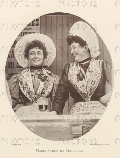 Portrait of two waffle sellers in Antwerp, Marchandes de gauffres, page, photomechanical print, photographer: Victor Selb, Antwerp, printer (person): Joseph Maes, c. 1890 - in or before 1894, printer (person), paper, collotype, photoprint, height 203 mm x width 172 mm, merchant, salesman - BB - women engaged in trade and commerce, waffles, Two women sit side by side inside an oval frame, both wearing pleated round hats and patterned shawls over high-collared dresses; the woman on the left faces forward with a slight smile while the woman on the right turns toward her with a broader smile, and both have short curly hair and earrings. A table edge is visible in front of them with a glass placed on it, and the background shows vertical wooden panels. The image is sepia-toned.