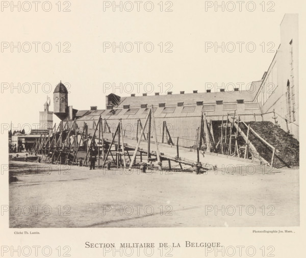 Wooden bridge in the military section of the world exhibition in Antwerp, Section Militaire de la Belgique, page, photomechanical print, photographer: Th. Lantin, Antwerp, printer (person): Joseph Maes, 1894, printer (person), paper, collotype, photoprint, height 160 mm x width 219 mm, world fair, world exhibition, improvised bridge, temporary bridge, military engineering, A wide, sepia-toned scene showing an industrial waterfront with a long building featuring a sloped roof and multiple skylights, supported by a series of wooden scaffolds and angled beams along its length, a ramp or walkway leading up to the structure with chains and bollards at the base, a tall cylindrical tower with a domed top near the left side, additional smaller buildings and chimneys behind the tower, a pile of material or vegetation on a sloped conveyor on the right, several small figures near the scaffolding, and an expanse of open ground in the foreground.