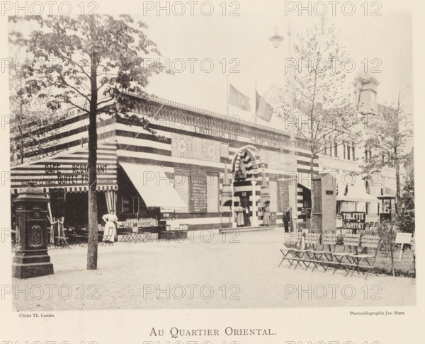 At the Oriental Quarter, Exterior of the Oriental section of the World Exhibition in Antwerp, page, photomechanical print, photographer: Th. Lantin, Antwerp, printer (person): Joseph Maes, 1894, printer (person), paper, collotype, photogravure, height 160 mm x width 219 mm, façade (of house or building), world fair, world exhibition, A wide exterior scene shows a striped building facade with an ornate arched entrance flanked by columns and decorative panels, flags flying above the roofline, and patterned awnings extending from the front; several people stand near the entrance and a woman in a dress stands by a cluster of small tables and chairs on the left; trees with leaves frame parts of the scene and a row of folding chairs lines the right foreground; a freestanding ornamental structure stands near the left edge and a tall lamp hangs above the street.