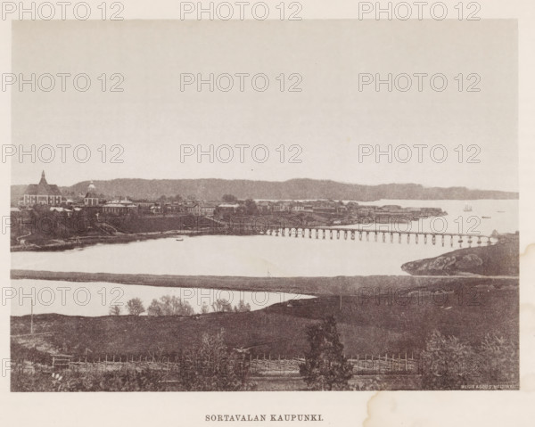 Sortavalan Kaupunki, View of Sortavala, page, photomechanical print, photographer: Impi Backman, Sortavala, cliché maker: anonymous, printer (person): Weilin & Göös, c. 1888 - c. 1893, printer (person), paper, autotypie, height 163 mm x width 224 mm, Print two pages for page 1., city-view in general, 'veduta', landscapes with waters, waterscapes, seascapes (in the temperate zone), bridge, A wide sepia-toned panorama shows a shoreline town across a calm body of water with a long wooden pier stretching from the right toward the center; buildings with varied rooflines and a few domed and spired structures sit along the distant shore backed by a low ridge of land, and a small sailboat is visible on the water; in the foreground there are grassy and rocky fields with scattered trees and simple wooden fences, and a strip of land or sandbar runs parallel to the nearer shore.
