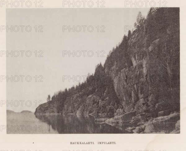 Rocks by a water, Haukkalahti. impilahti, page, photomechanical print, photographer: Impi Backman, cliché maker: anonymous, printer (person): Weilin & Göös, c. 1888 - c. 1893, printer (person), paper, autotypie, height 153 mm x width 212 mm, trees, landscapes with waters, waterscapes, seascapes (in the temperate zone), rocks, Karelia, A monochrome photograph showing a rocky shoreline with steep cliffs rising from calm water, trees growing along the cliff tops and scattered on rocky ledges, reflections of the rocks and trees visible in the smooth surface of the water, and distant low hills fading into the background under a pale sky.