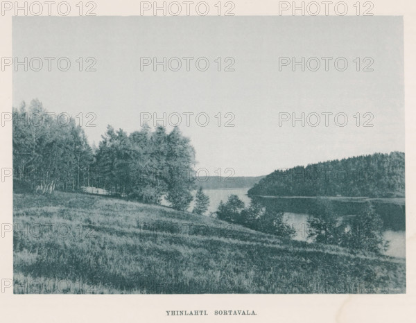 View of a river in a landscape with trees, Yhinlahti Sortavalan, page, photomechanical print, photographer: Impi Backman, cliché maker: anonymous, printer (person): Weilin & Göös, c. 1888 - c. 1893, printer (person), paper, autotypie, height 164 mm x width 230 mm, trees, landscapes with waters, waterscapes, seascapes (in the temperate zone), Karelia, A grainy landscape image in muted tones depicting a gently sloping grassy field in the foreground, a stand of leafy trees along the left edge, and a body of water extending toward a wooded shoreline on the right, with distant tree-covered hills visible across the water and a wide pale sky occupying the upper portion of the scene.
