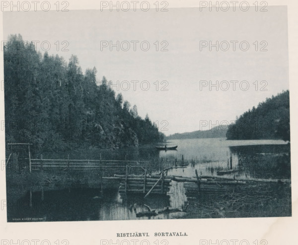 Ristijärvi . Sortavalan, View of a canoe on water, page, photomechanical print, photographer: Impi Backman, cliché maker: anonymous, printer (person): Weilin & Göös, c. 1888 - c. 1893, printer (person), paper, autotypie, height 156 mm x width 211 mm, rowing-boat, canoe, landscapes with waters, waterscapes, seascapes (in the temperate zone), rocks, Karelia, A lakeshore scene with calm water stretching toward a distant tree-lined horizon, steep forested slopes on both sides, a small rowboat with a person seated in it near the center of the water, a foreground wooden structure made of logs and rails extending into the water, reflected shapes on the water surface, reeds and silhouetted vegetation at the water’s edge, and a pale sky above.