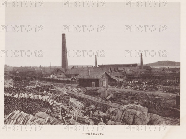 Chimneys near buildings and stacks of wood, Pitkäranta, page, photomechanical print, photographer: Impi Backman, cliché maker: anonymous, printer (person): Weilin & Göös, c. 1888 - c. 1893, printer (person), paper, autotypie, height 157 mm x width 226 mm, civic architecture, edifices, dwellings, chimney, industries, plant material, A sepia-toned landscape showing a shoreline area with rocky foreground and scattered piles of cut logs and planks, a cluster of simple wooden houses and outbuildings with pitched roofs set behind the rocky area, a larger industrial complex behind the houses with long low buildings and several tall narrow smokestacks rising into the pale sky, distant low hills and water visible along the horizon, and an expansive empty sky occupying the upper portion of the image.