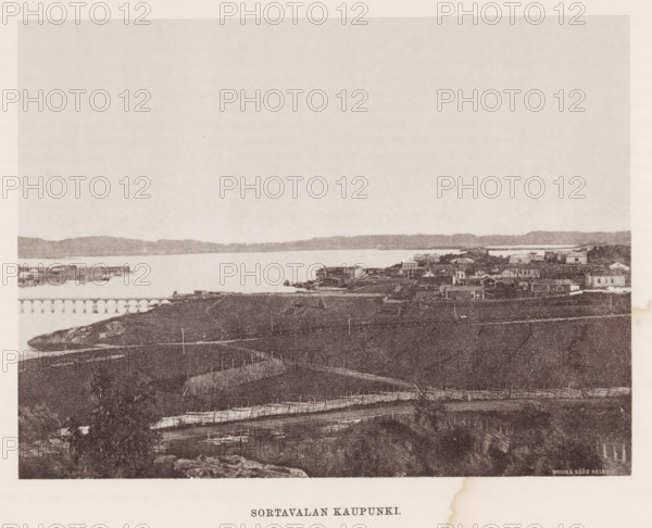 View of Sortavala, Sortavalan Kaupunki, page, photomechanical print, photographer: Impi Backman, Sortavala, cliché maker: anonymous, printer (person): Weilin & Göös, c. 1888 - c. 1893, printer (person), paper, autotypie, height 157 mm x width 210 mm, city-view in general, 'veduta', landscapes with waters, waterscapes, seascapes (in the temperate zone), bridge, A wide sepia-toned shoreline view with a calm body of water stretching across the middle, a distant low ridge on the far side, and a small cluster of buildings and houses along the near shore including piers and docks; in the foreground there are fenced plots and narrow paths running across a grassy, uneven landscape with rocks and scattered vegetation, and a faint sky occupying the upper portion of the image.