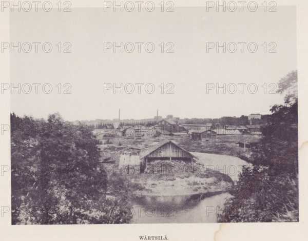 Wood storage facility in a landscape, Wärtsilä, page, photomechanical print, photographer: Impi Backman, cliché maker: anonymous, printer (person): Weilin & Göös, c. 1888 - c. 1893, printer (person), paper, autotypie, height 164 mm x width 217 mm, landscapes with waters, waterscapes, seascapes (in the temperate zone), civic architecture, edifices, dwellings, Pohjois-Karjala, Värtsilä, Vyartsilya, A grainy monochrome scene showing a riverside with several wooden buildings and sheds along the bank, a larger barn-like structure near the center with stacks of firewood visible under its overhang, a narrow waterway curving past the foreground reflecting surrounding shapes, dense trees and foliage framing both sides of the image, a low distant skyline of more industrial and residential buildings and two tall slender chimneys rising above the roofs, and a wide pale sky occupying the upper portion of the picture.
