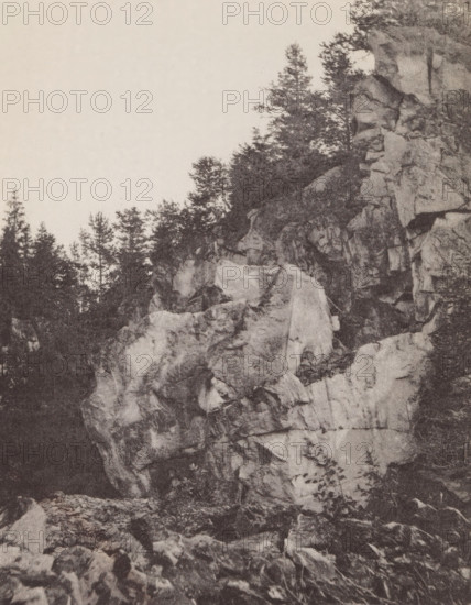 Ruskaelan Marmorilouhos, Rocks in Ruskeala, page, photomechanical print, photographer: Impi Backman, cliché maker: anonymous, printer (person): Weilin & Göös, c. 1888 - c. 1893, printer (person), paper, autotypie, height 215 mm x width 167 mm, rocks, Sortavala, A tall rocky cliff face occupies the right side of the image, showing layered rough stone with visible cracks and ledges, while trees and dense foliage grow along the top edge and in the background on the left; the foreground contains scattered rocks and boulders at the cliff base, and the sky above is clear and light in tone.