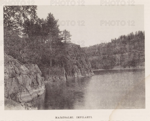 View of a river, Majatsalmi Impilahti, page, photomechanical print, photographer: Impi Backman, cliché maker: anonymous, printer (person): Weilin & Göös, c. 1888 - c. 1893, printer (person), paper, autotypie, height 152 mm x width 206 mm, trees, river, rocks, Karelia, A wide monochrome scene showing a rocky shoreline with steep cliffs and rough textured rock faces rising from calm water, trees and dense forest vegetation covering the cliff tops and extending into the background, a smooth expanse of water reflecting subtle ripples along the shore, and a pale sky above with no visible clouds.