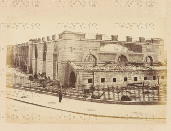 Grenier d'Abondance - vue extérieure, View of a destroyed building, photograph, page, 1871, photographer, paper, albumen print, albumendruk, height 101 mm x width 151 mm, ruin of a dwelling, house, castle, palace, Paris, A sepia-toned photograph of a large stone building with a long facade featuring arched openings and rectangular blocks of masonry, a lower section with small square openings and a roof edge showing debris, upper walls with arched window openings and some metal grilles, and a parapet with short square pillars and railings; in front of the building a fenced yard contains scattered rubble and large cylindrical objects, the fence running along a paved street where a solitary figure wearing a long coat and hat stands near the curb.