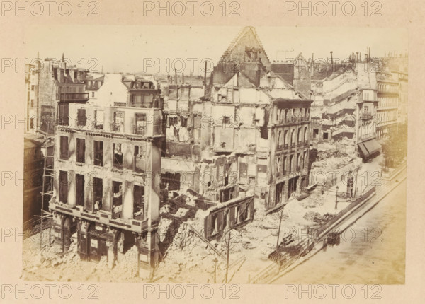 View of a destroyed Théâtre de la Porte Saint-Martin, Théatre de la Porte St Martin, photograph, page, Paris, 1871, photographer, paper, albumen print, height 101 mm x width 151 mm, ruin of a dwelling, house, castle, theater (building), Théâtre de la Porte Saint-Martin, A sepia-toned aerial view shows a row of heavily damaged multi-story buildings with exposed interiors, missing walls, collapsed floors, rubble piled at street level, and jagged rooflines; scaffolding and wooden beams lean against some facades, a tram or rail track runs along a nearby street where a small vehicle and carts are visible, and bare trees and lampposts line the sidewalk beside debris-strewn pavement.